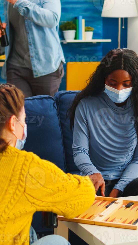 African woman competing with friends playing backgammon wearing face mask keeping social distancing during social pandemic with covid19 drinking beer in living room. Enjoying board games in outbreak photo