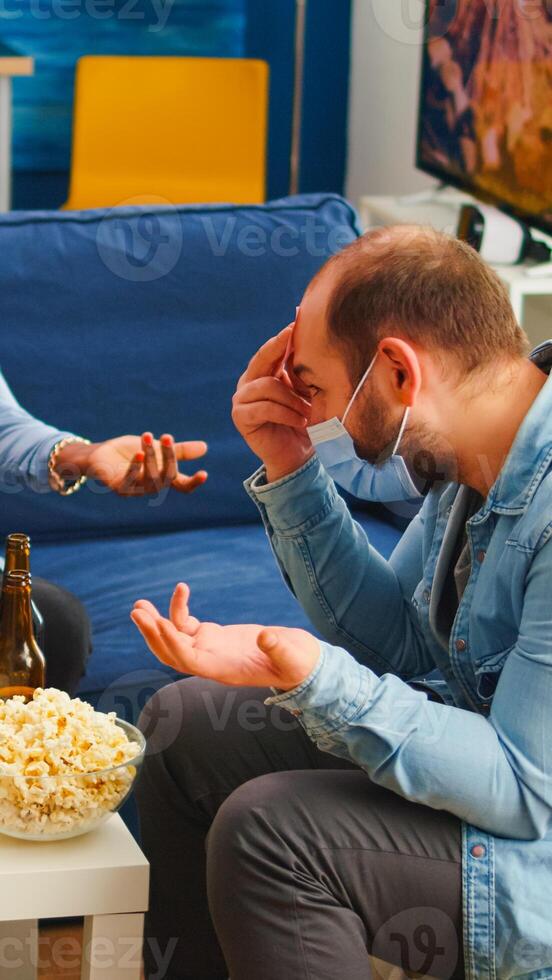 Thoughtful multiethnic friends with sticky notes on foreheads playing name game miming gesturing wearing face mask keeping social distancing preventing covid19 spread. People enjoying bottles of beer photo