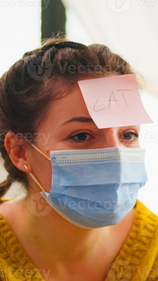 Happy woman gesturing playing name game with multiethnic friends having sticky notes on forehead wearing face mask keeping social distancing. Diverse group of people having fun at new normal party photo