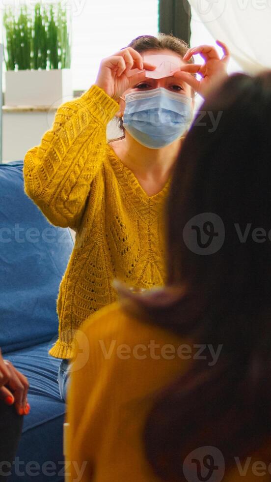 Happy woman with sticky note on forehead playing name game with multiethnic friends keeping social distancing drinking beer in home living room due to social pandemic. People having fun in outbreak photo