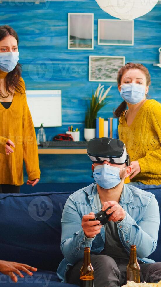 Group of multi ethnic friends having fun playing games using virtual reality headset and joystick wearing face mask keeping social distancing. Diverse people enjoying at new normal party photo