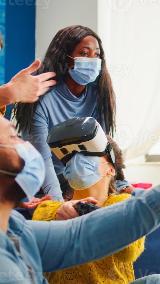 Friends supporting woman during game playing with virtual reality goggles wearing protection mask agaist covid19 virus. Diverse people having fun, laughing, drinking beer at new normal party photo
