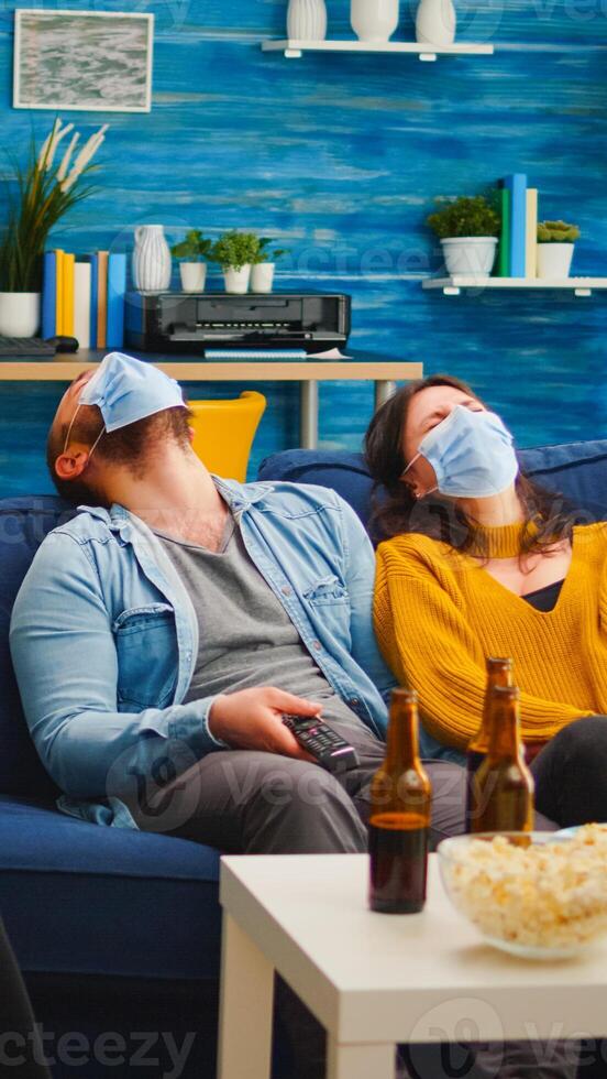 Group of multi ethnic friends watching comedy show on tv laughing wearing face mask to prevent infection with covid 19, during global pandemic having fun sitting on couch keeping social distance photo