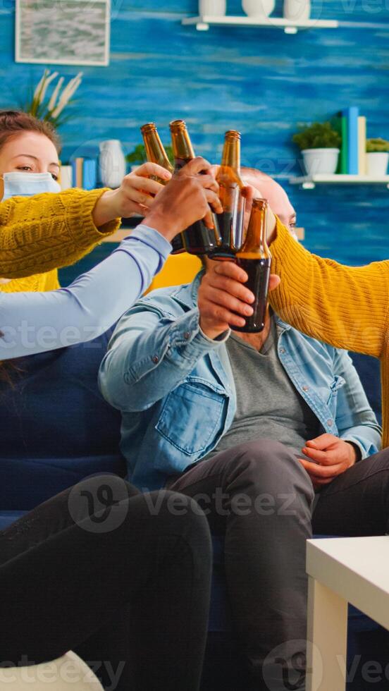 Diverse group of friends watching TV together comedy sitcom eating snacks and drinking beer, clinking bottles and having fun at new normal party, wearing protection face mask against covid 19 virus photo