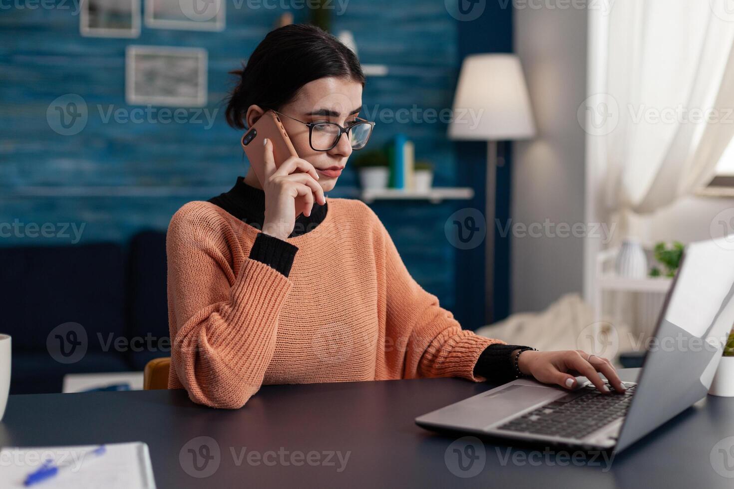 Student at home talking on the phone while using laptop. Programmer with glasses working from home using smartphone and browsing computer. Focused freelancer using technology for remote work. photo