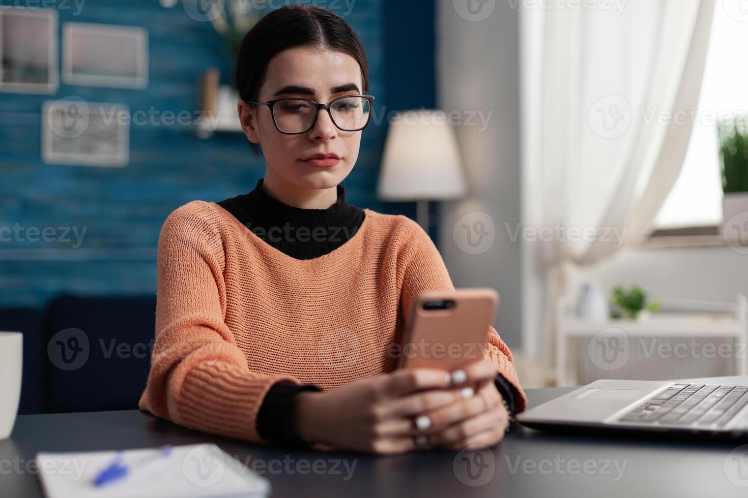 Student with glasses reading email on smartphone. Freelancer holding smart phone looking at content on screen. Influencer sitting at desk browsing social media on tochscreen smart phone. photo