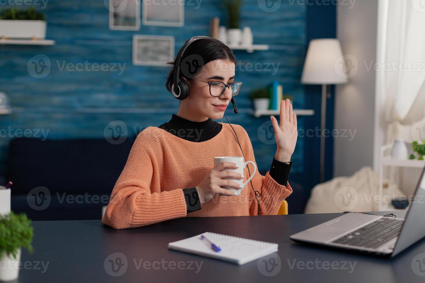Student with headphones waving hello in laptop videocall. College girl with glasses holding cup attendig online call lecture. Programmer raising hand in remote teleconference course. photo