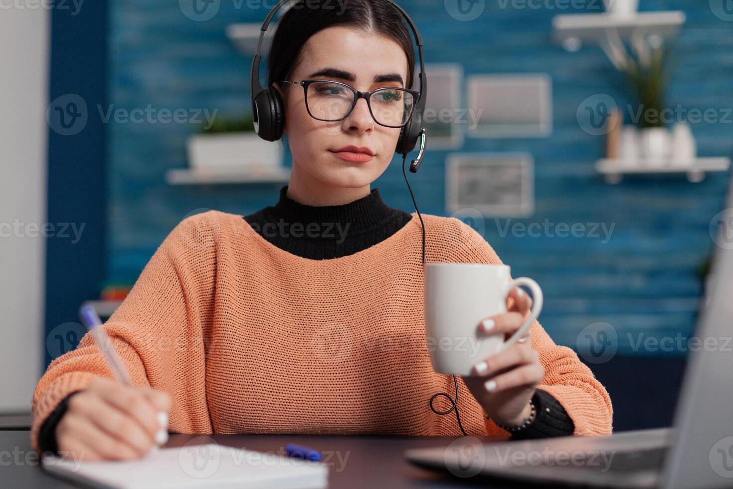 Student in online class holding cup looking at laptop screen and taking notes. Woman with headphones writing on notebook listening to university lecture. College girl with glasses studying remote. photo