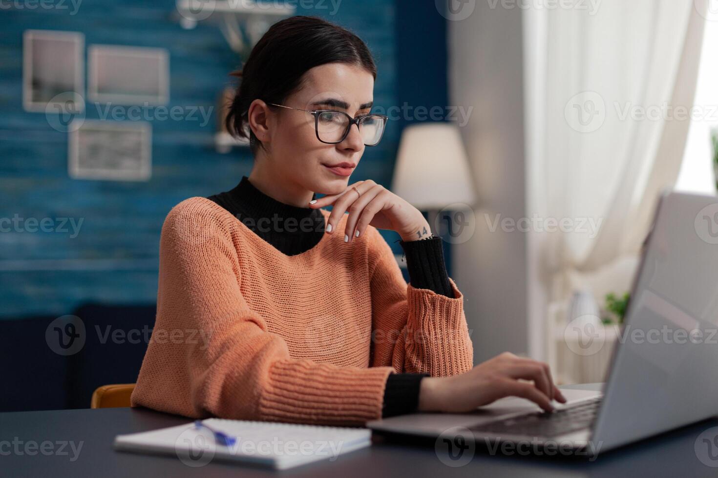 Confident student studying remote looking at laptop screen. Content creator with glasses casually typing sitting at home desk. College girl browsing the internet. Freelancer working from home sending email. photo