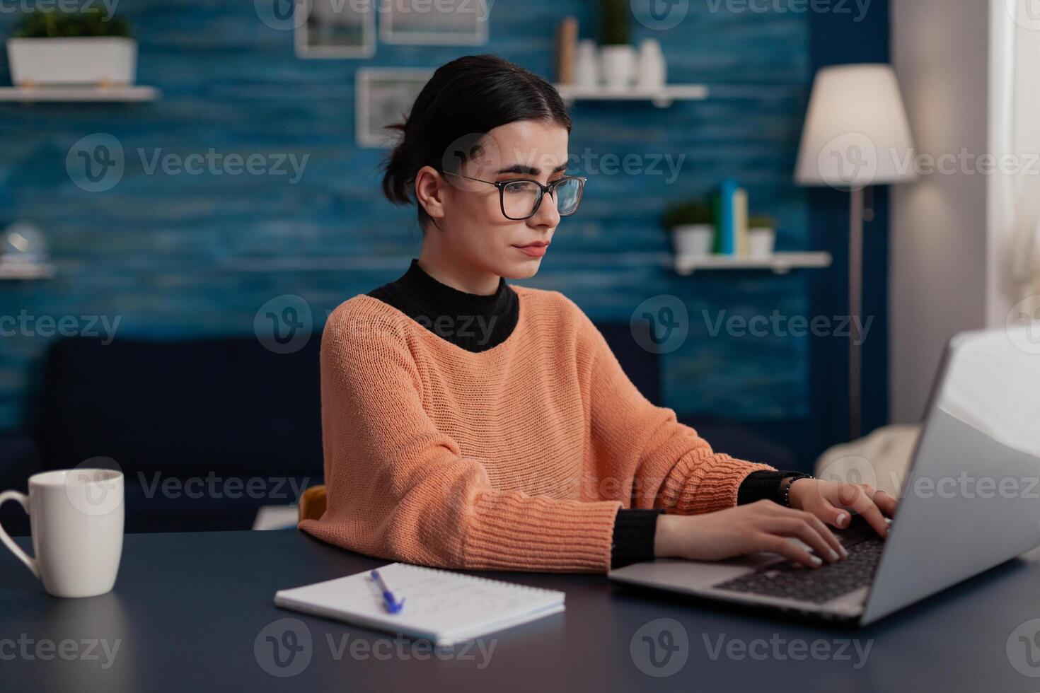 Student with notebook studying remote looking at laptop screen. Influencer with glasses typing on keyboard sitting at home desk. College girl browsing the internet. Freelancer sending email. photo
