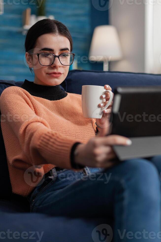 Student with glasses on couch holding cup looking at tablet display. Influencer on sofa in videocall conference while drinking coffee or tea. Content creator streaming on digital device. photo