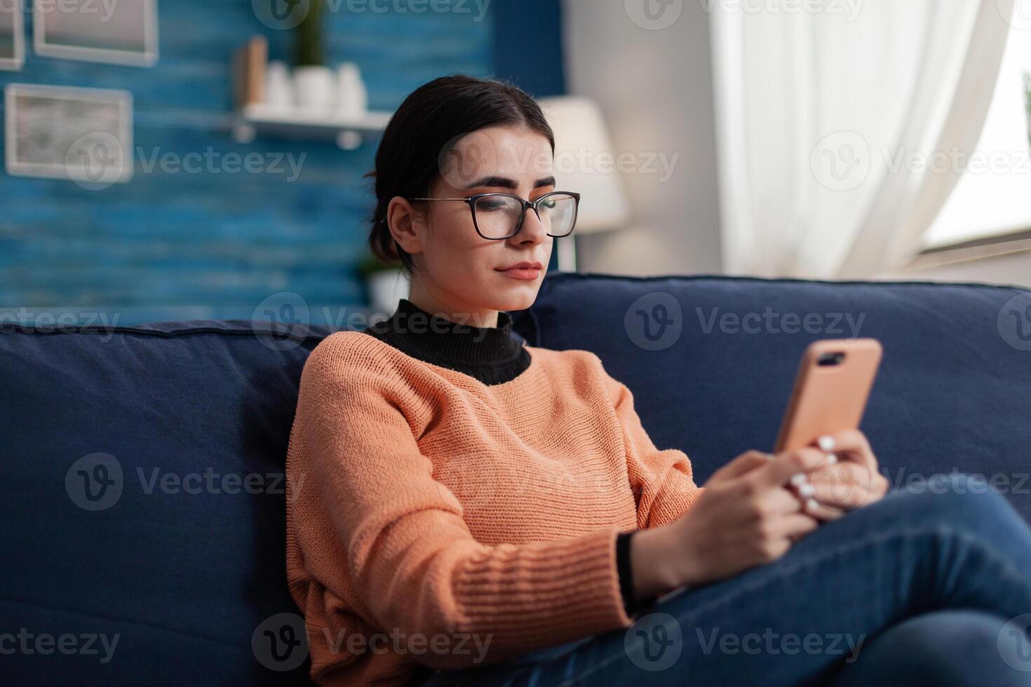 Student relaxing on couch holding smartphone and looking at screen. Content creator with glasses on sofa reading content on mobile phone. Digital influencer checking email on tochscreen device. photo
