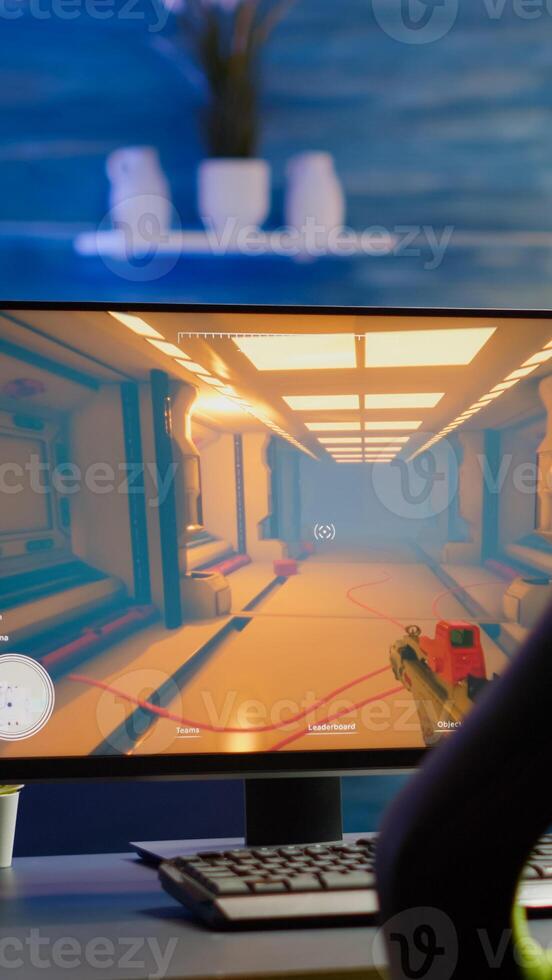 RGB powerful personal computer gamer rig with first-person shooter game on screen. Monitor stands on table in home gaming room. Cozy room with modern design is lit with warm and neon light. photo
