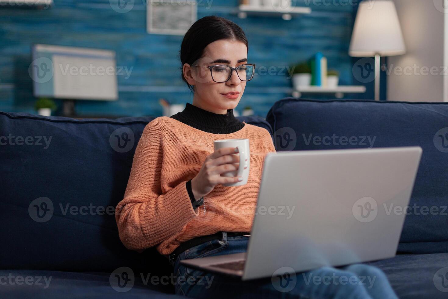 Student sitting on sofa holding cup and laptop. Freelance programmer on couch with glasses working from home in living room. Content creator browsing on social media using digital device. photo
