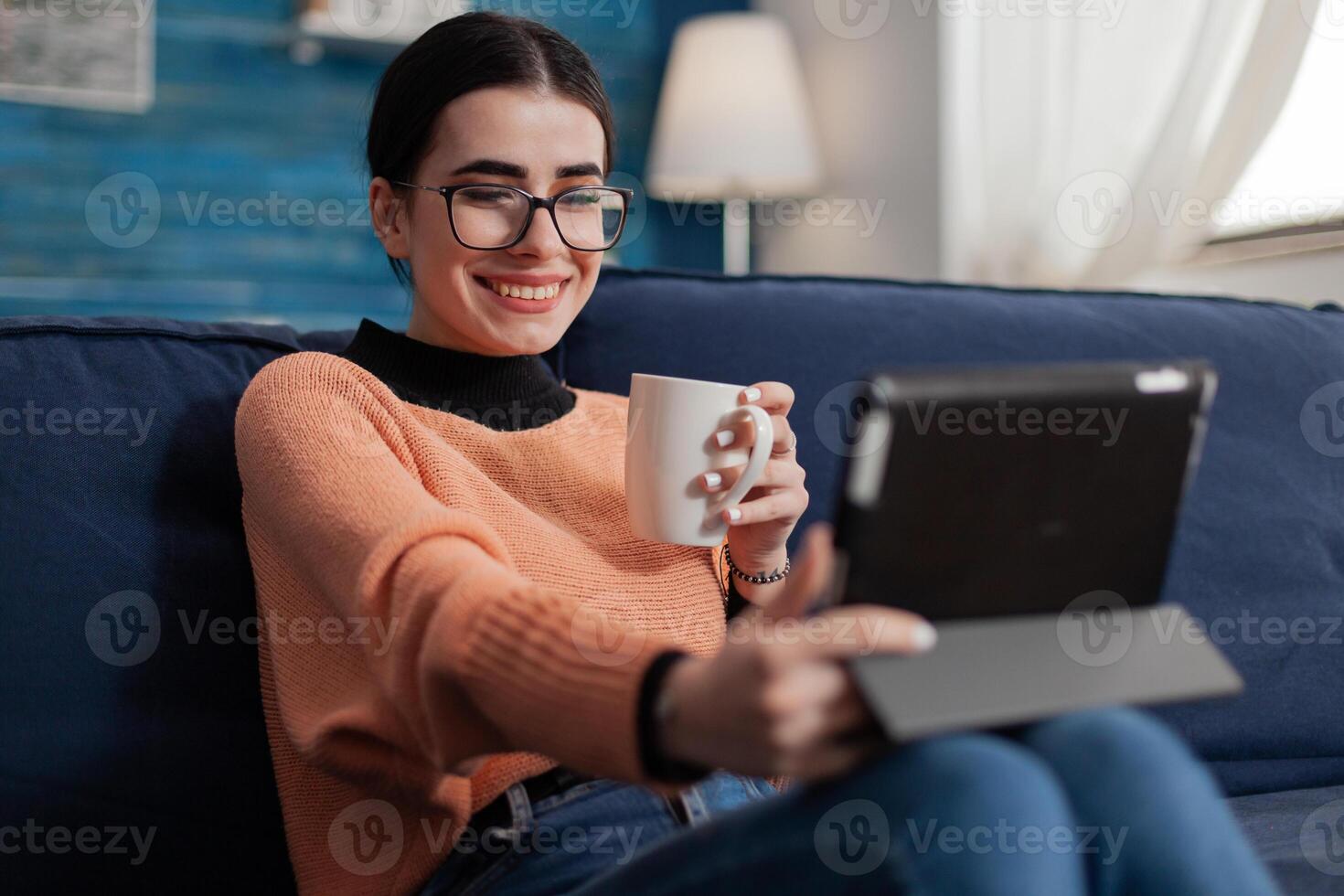 Smiling student on sofa casually talking in videocall using tablet. Content creator with glasses on couch watching on social media. Digital influencer relaxing with cup of coffee or tea. photo