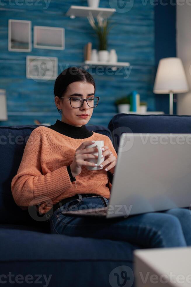 Content creator on couch with laptop on lap holding cup. Student with glasses sitting on sofa learning remote from home in living room. Influencer relaxing looking at screen. photo