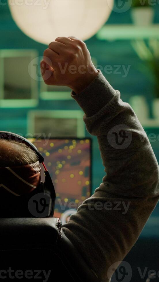 Back shot of happy gamer winning first person shooter game playing online on RGB powerful personal computer raising hands. Pro cyber performing during virtual tournament using technology network photo