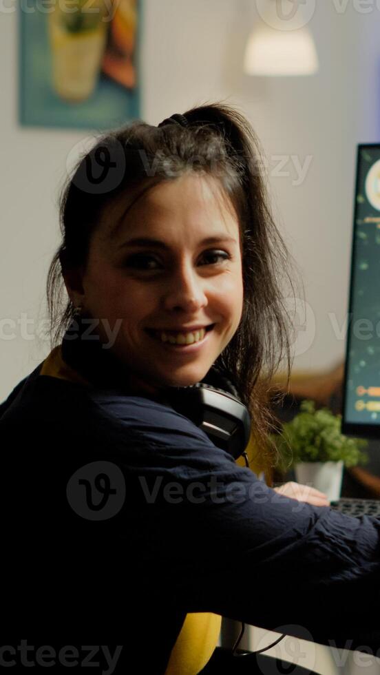 Woman gamer with headset looking at camera smiling and playing virtual shooter game championship in cyberspace, esports player performing on powerful computer in stylish room during gaming tournament photo