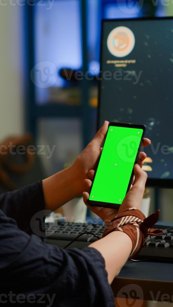 Professional cyber gamer looking at phone with greenscreen, chroma key, mock up desktop isolated display playing games on powerful computer. Player streaming shooter games wearing headset photo