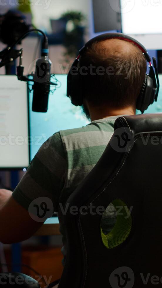 Back shot of streamer man playing on powerful computer shooter game for tournament, talking with multiple players into headphones. Pro game r using professional streaming equipment photo
