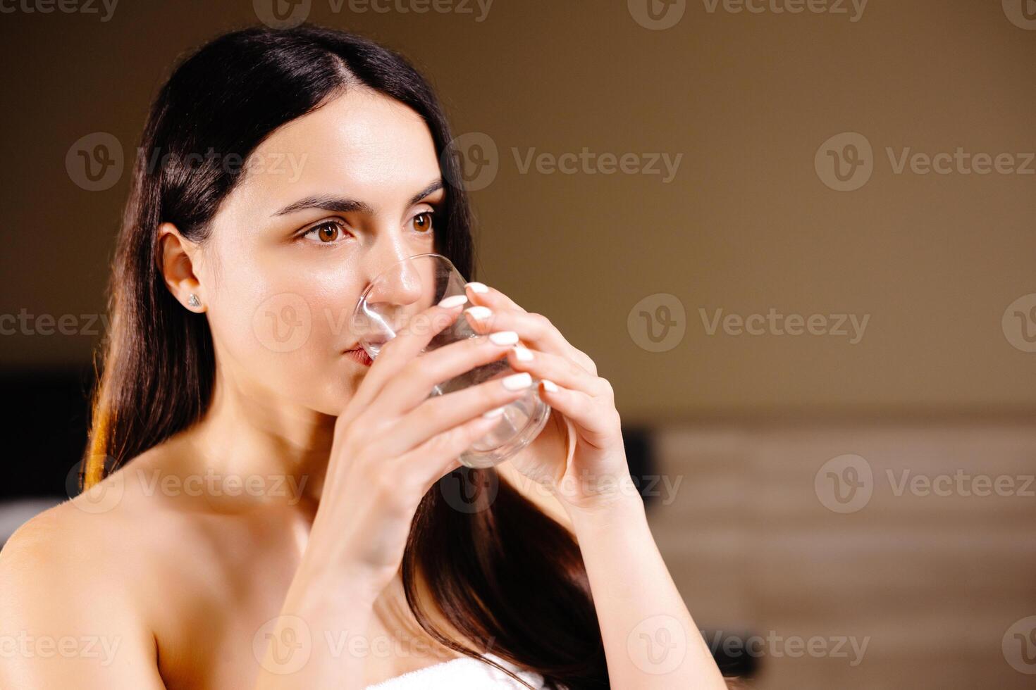 Pretty brunette drinking water standing near bed at home in the bedroom. photo