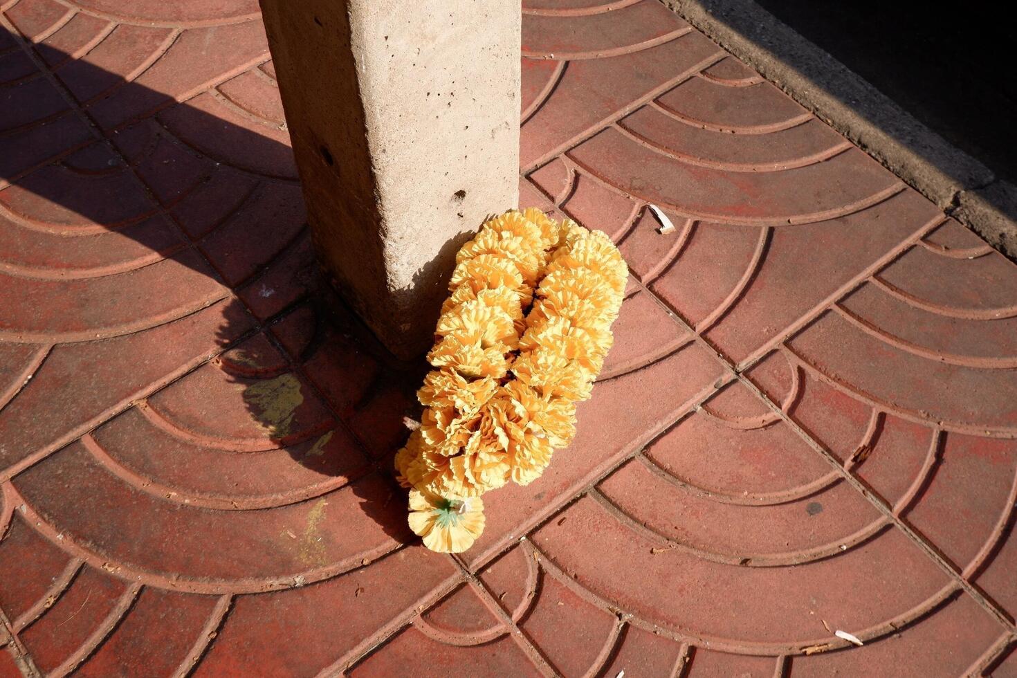 A garland of yellow marigold flowers placed at the base of a concrete pillar on a red-tiled sidewalk. photo