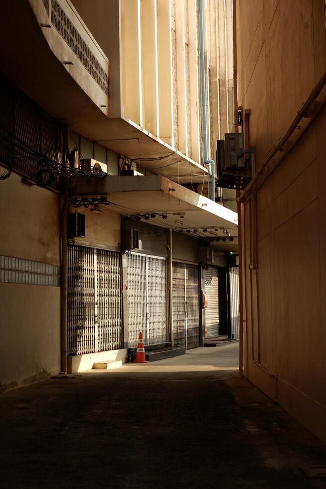 A narrow alleyway with closed metal gates on both sides. The sunlight filters through the buildings, casting dramatic shadows on the walls and floor. photo