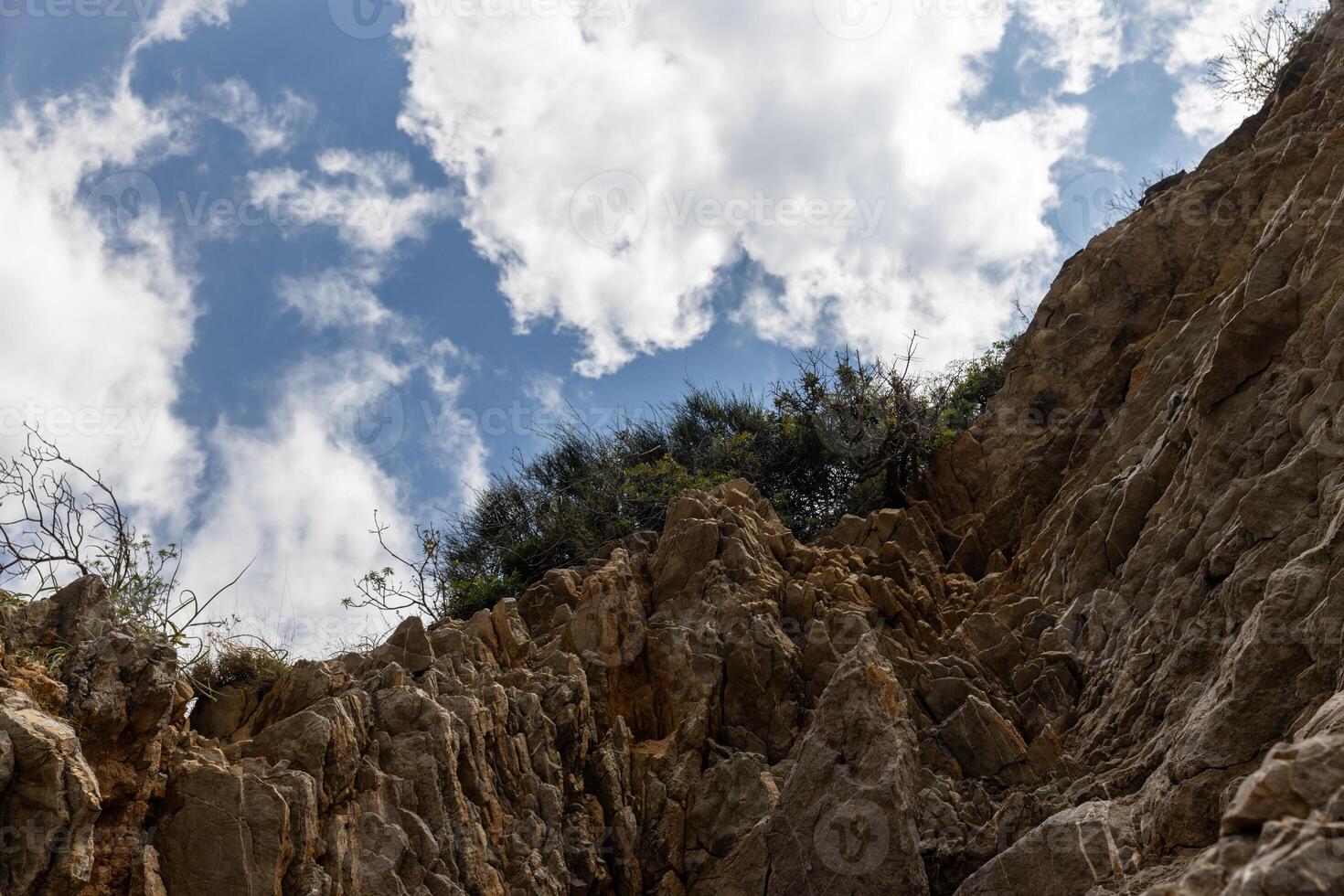 View from below of a rocky cliff with vegetation above against a blue sky and white clouds. Copy space.nature or abstract content, photo