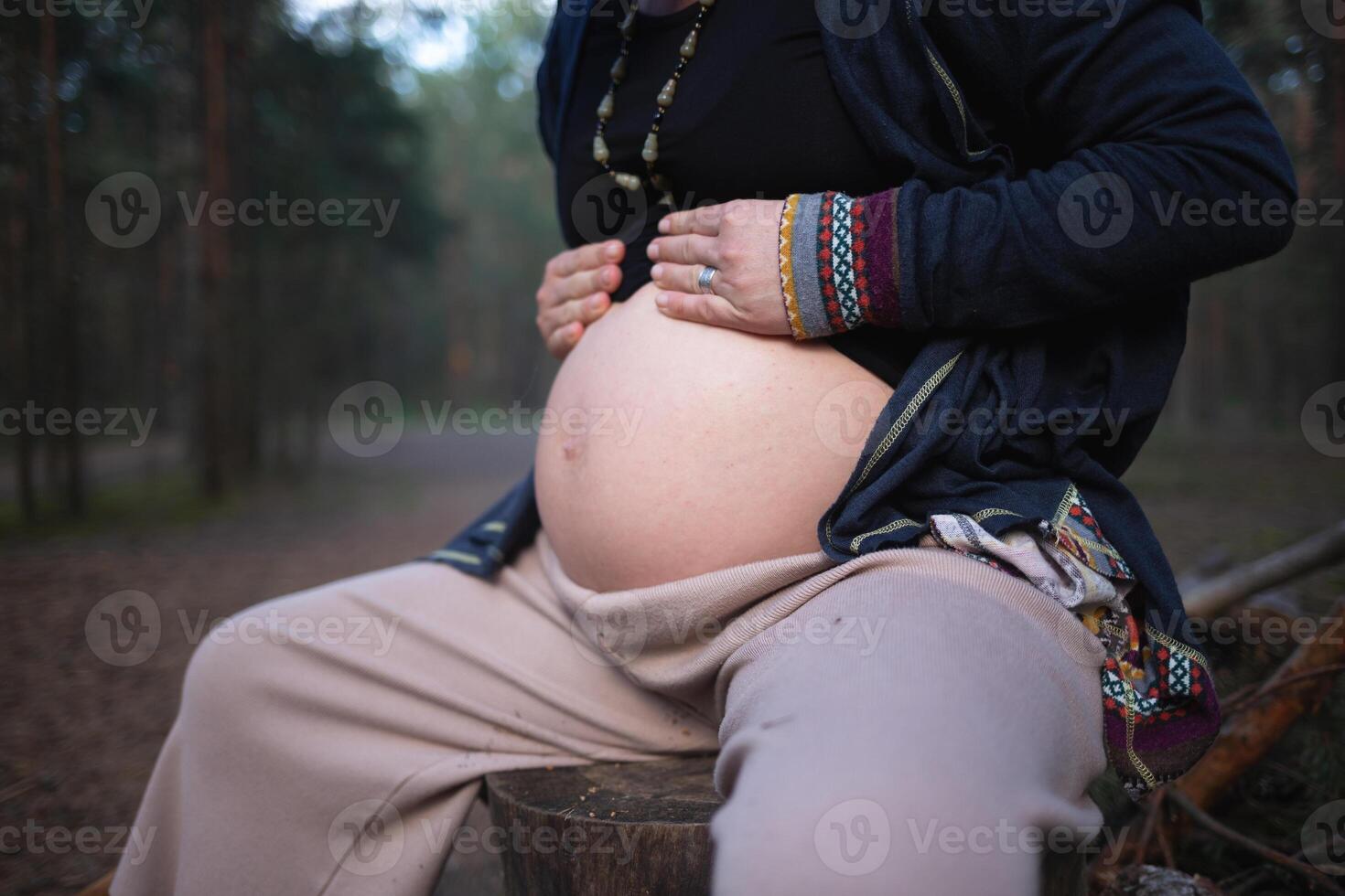 a pregnant woman sits on a log in the forest and hugs her tummy photo