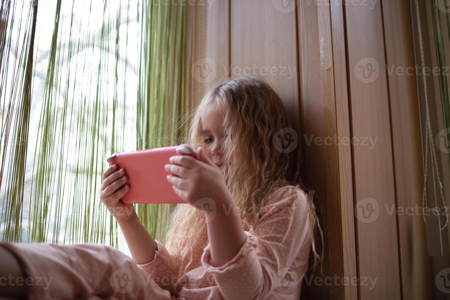 little cute girl plays a game console sitting on the windowsill photo