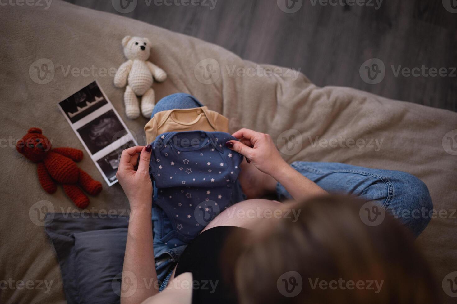 beautiful young pregnant woman sits on the bed in the bedroom and holds her baby's tiny clothes in her hands photo