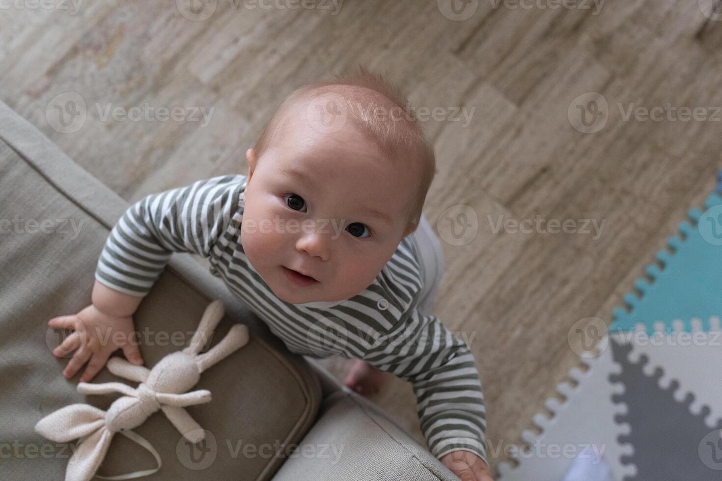 A curious baby in a striped onesie looks up, holding a toy rabbit with wonder photo