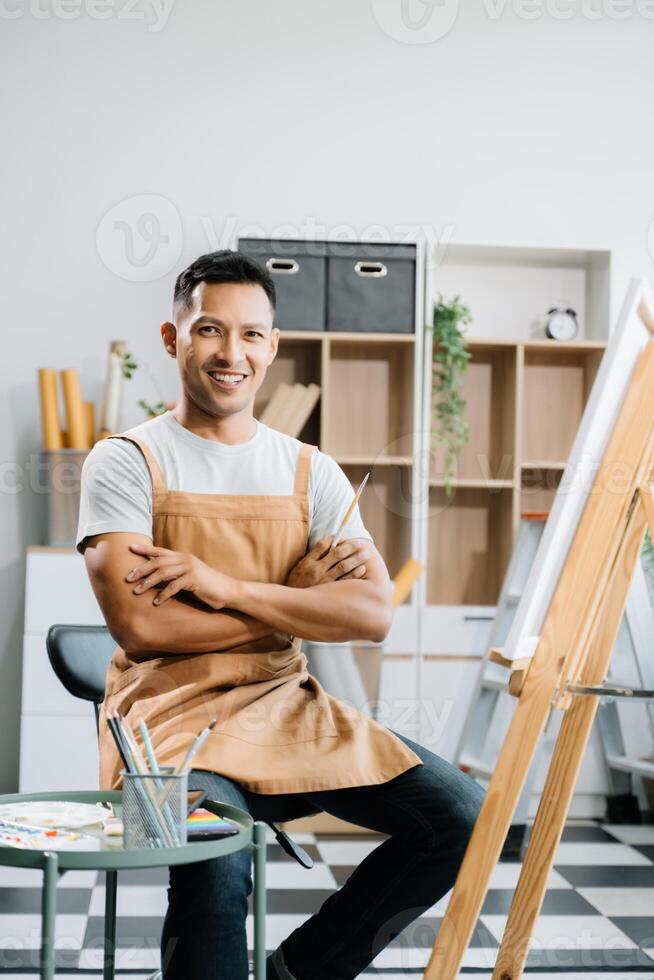 Asian male painter do artwork in art workshop, painting supplies, oil pastels, two canvas easel, creative space with paintbrush in art studio photo