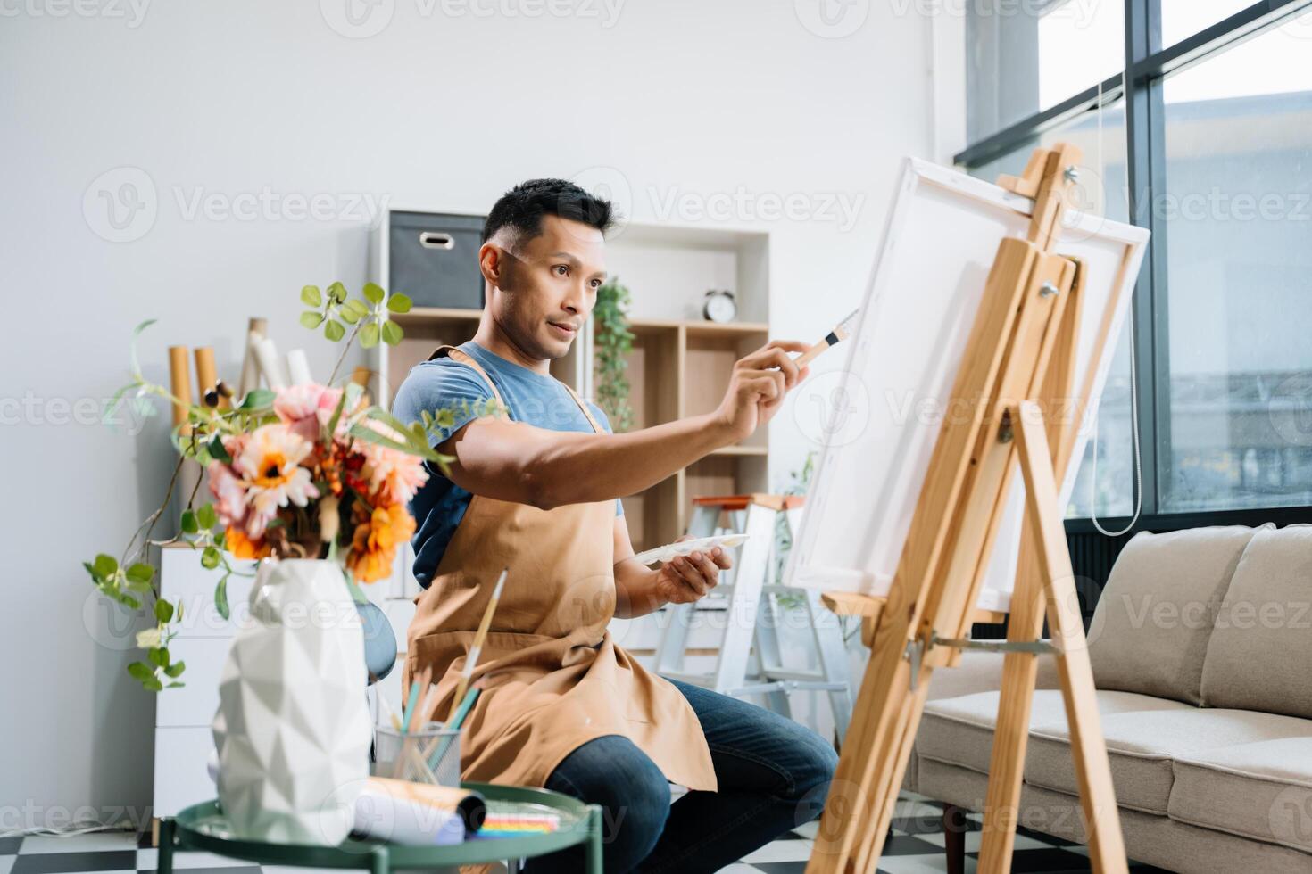Asian male painter do artwork in art workshop, painting supplies, oil pastels, two canvas easel, creative space with paintbrush in art studio photo