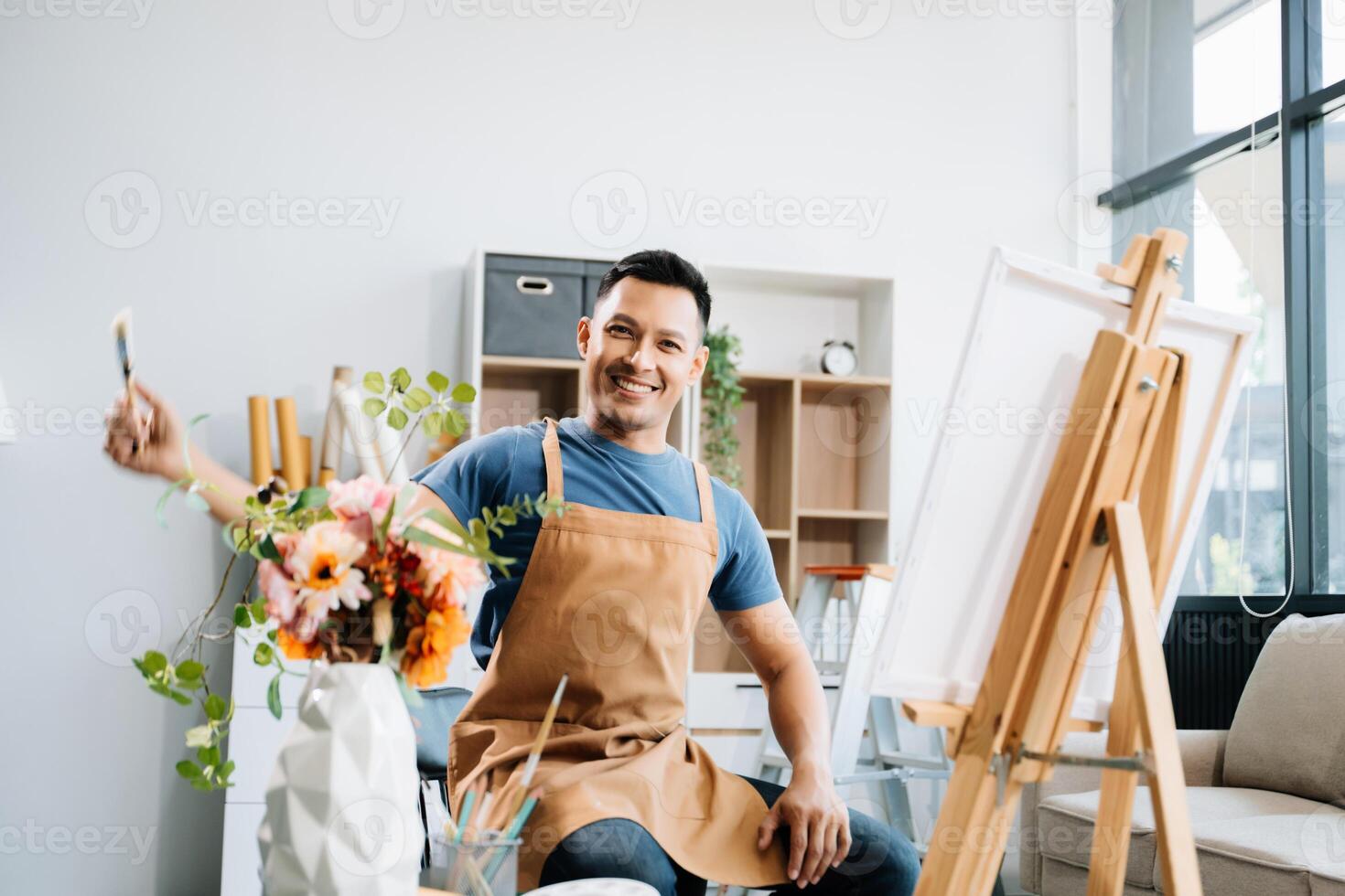 Asian male painter do artwork in art workshop, painting supplies, oil pastels, two canvas easel, creative space with paintbrush in art studio photo