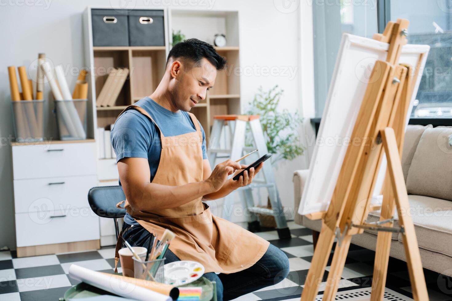 Asian male painter do artwork in art workshop, painting supplies, oil pastels, two canvas easel, creative space with paintbrush in art studio photo