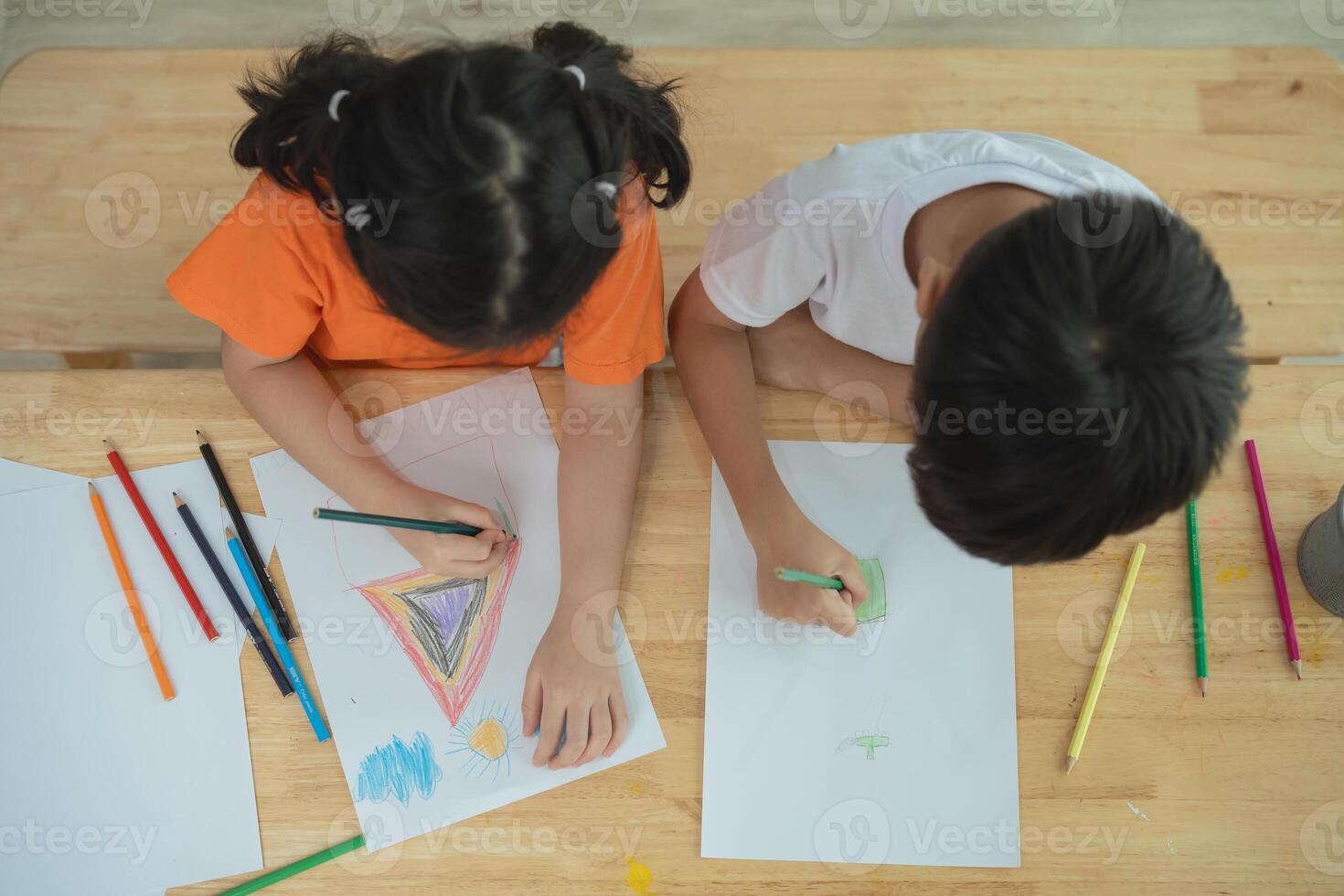 Two children drawing on a table with a variety of colored pencils. One child is drawing a triangle and the other is drawing a kite photo