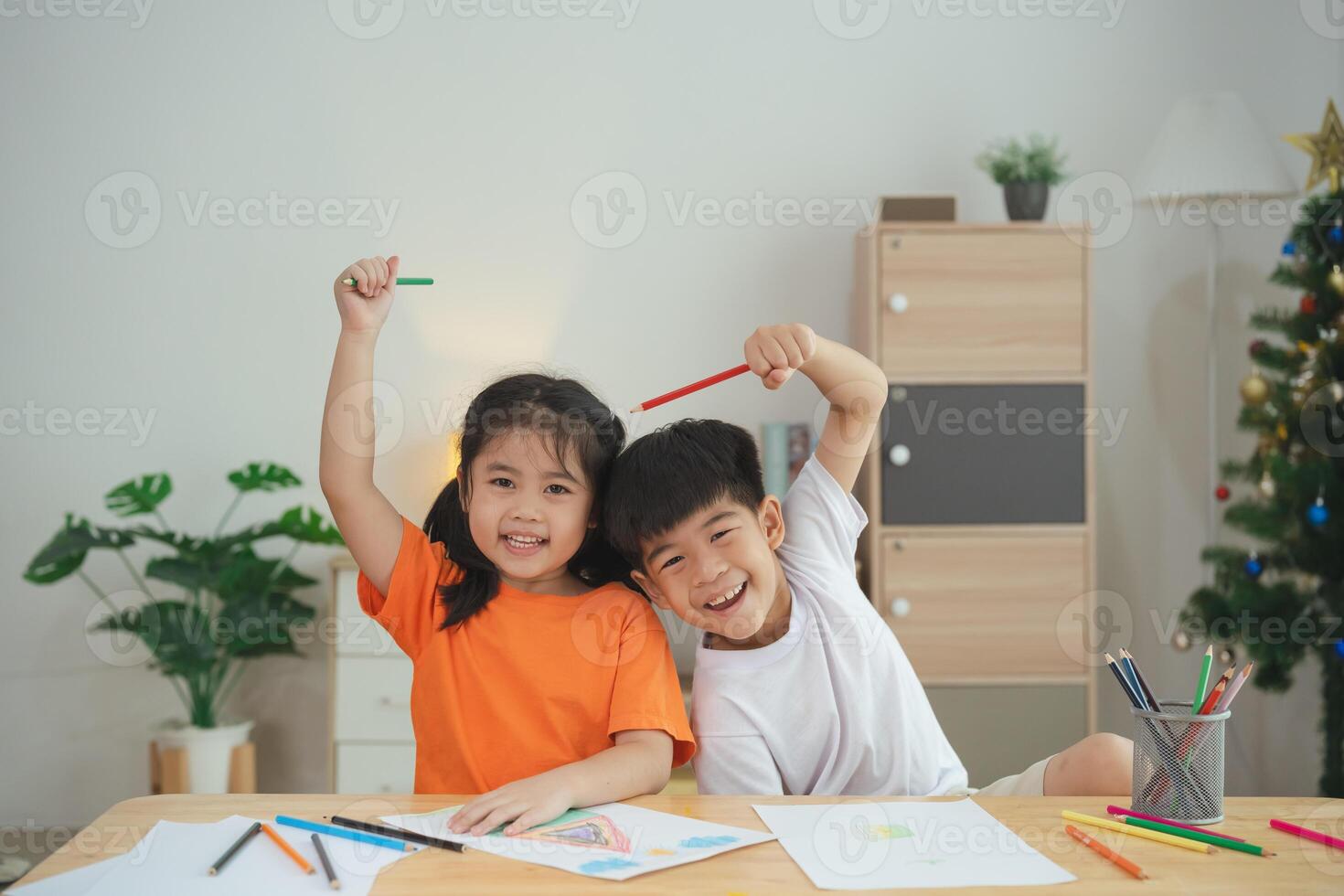 Two children are sitting at a table with crayons and drawing paper. One of the children is holding a pencil and the other is holding a crayon. They are both smiling and seem to be enjoying themselves photo