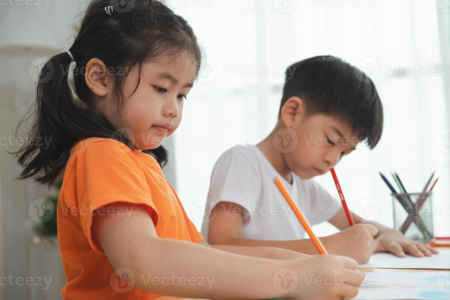 Two children are sitting at a table, drawing with crayons. One of the children is wearing an orange shirt photo