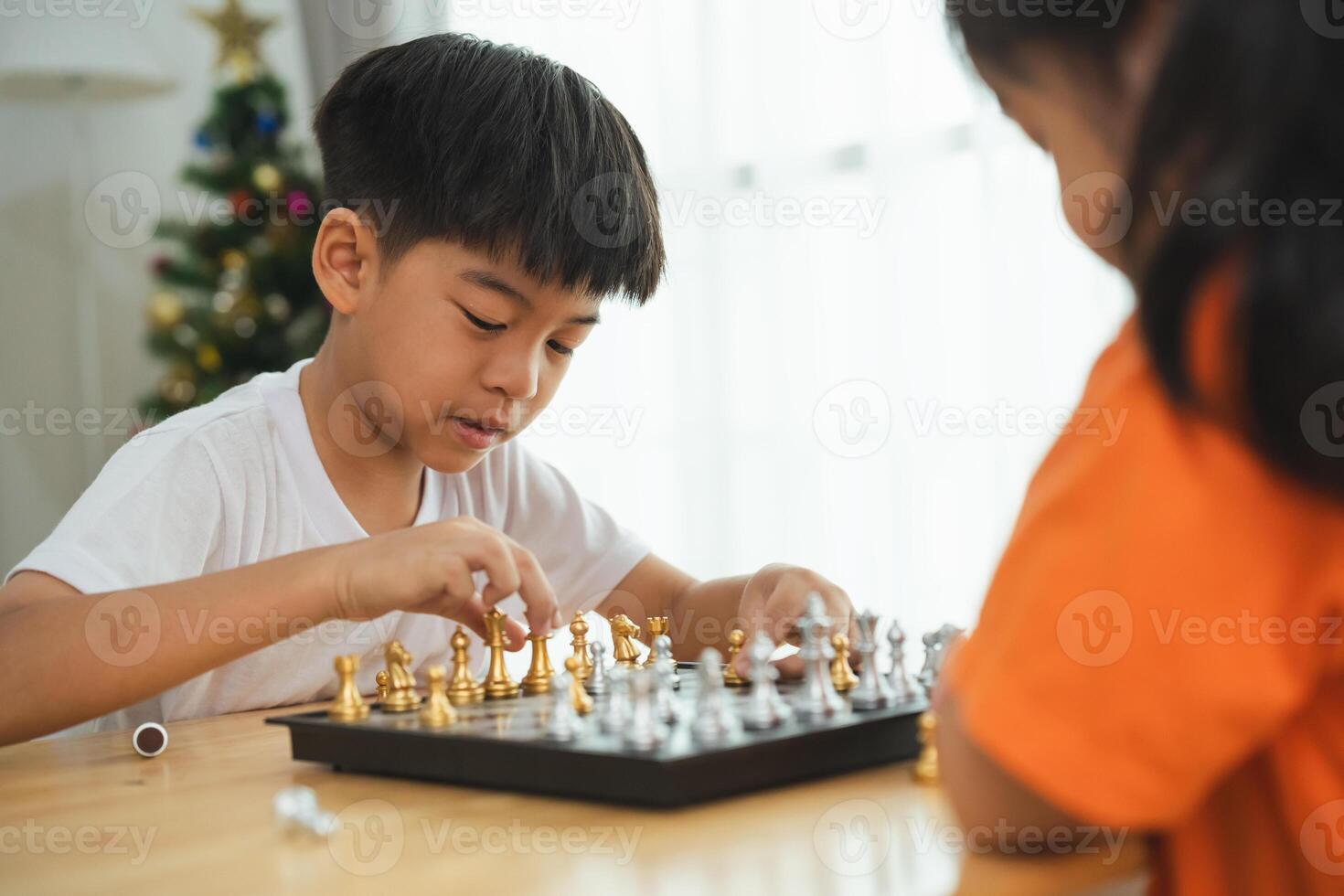 Two children playing a game of chess. One is smiling and the other is frowning. The boy in the white shirt is smiling and the girl in the orange shirt is frowning photo