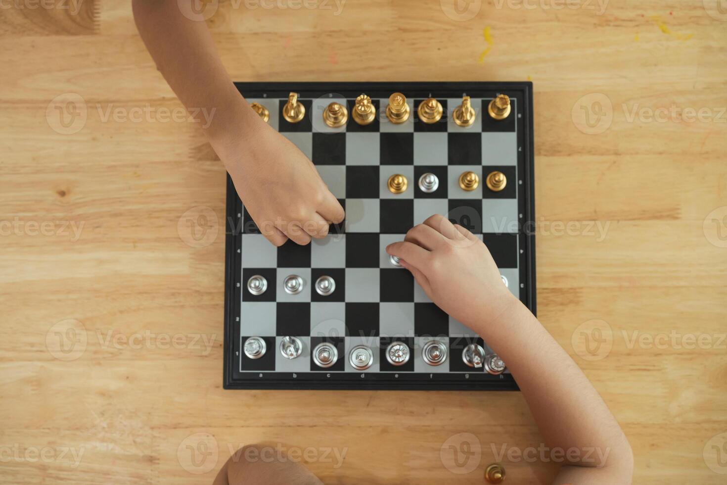 Two children playing a game of chess on a wooden table. One of the children is holding a piece in his hand photo
