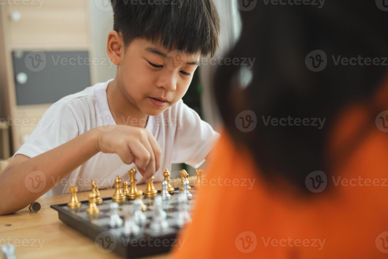 Two children playing a game of chess. One is smiling and the other is frowning. The boy in the white shirt is smiling and the girl in the orange shirt is frowning photo