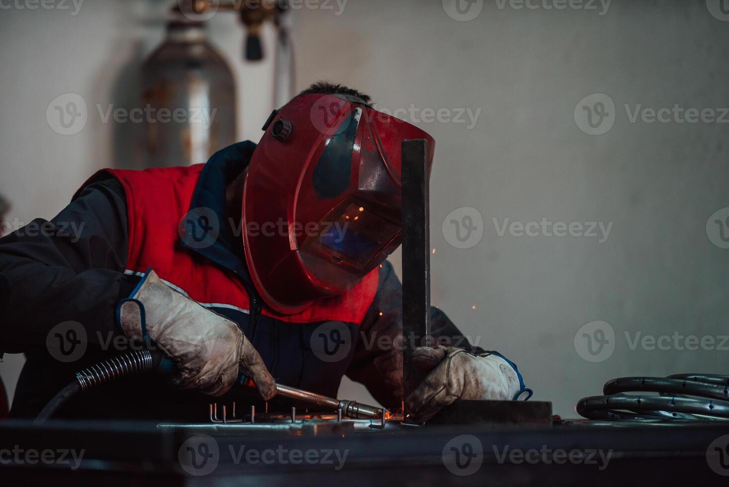 Profesional welder in protective uniform and mask welding metal pipe on the industrial table with other workers behind in the industrial workshop. photo