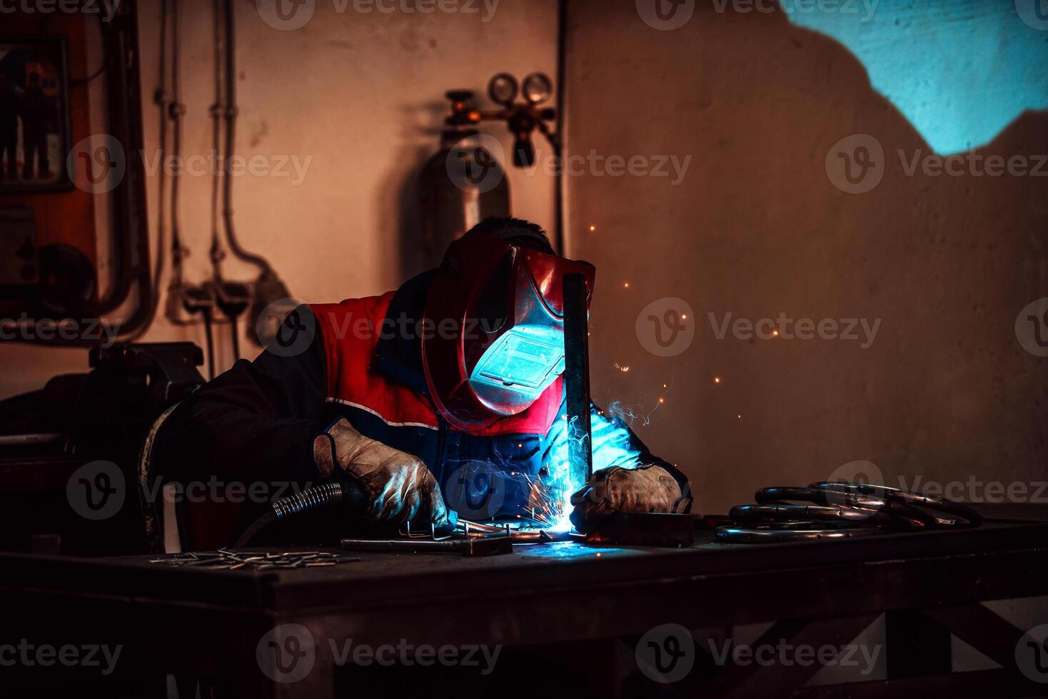 Profesional welder in protective uniform and mask welding metal pipe on the industrial table with other workers behind in the industrial workshop. photo