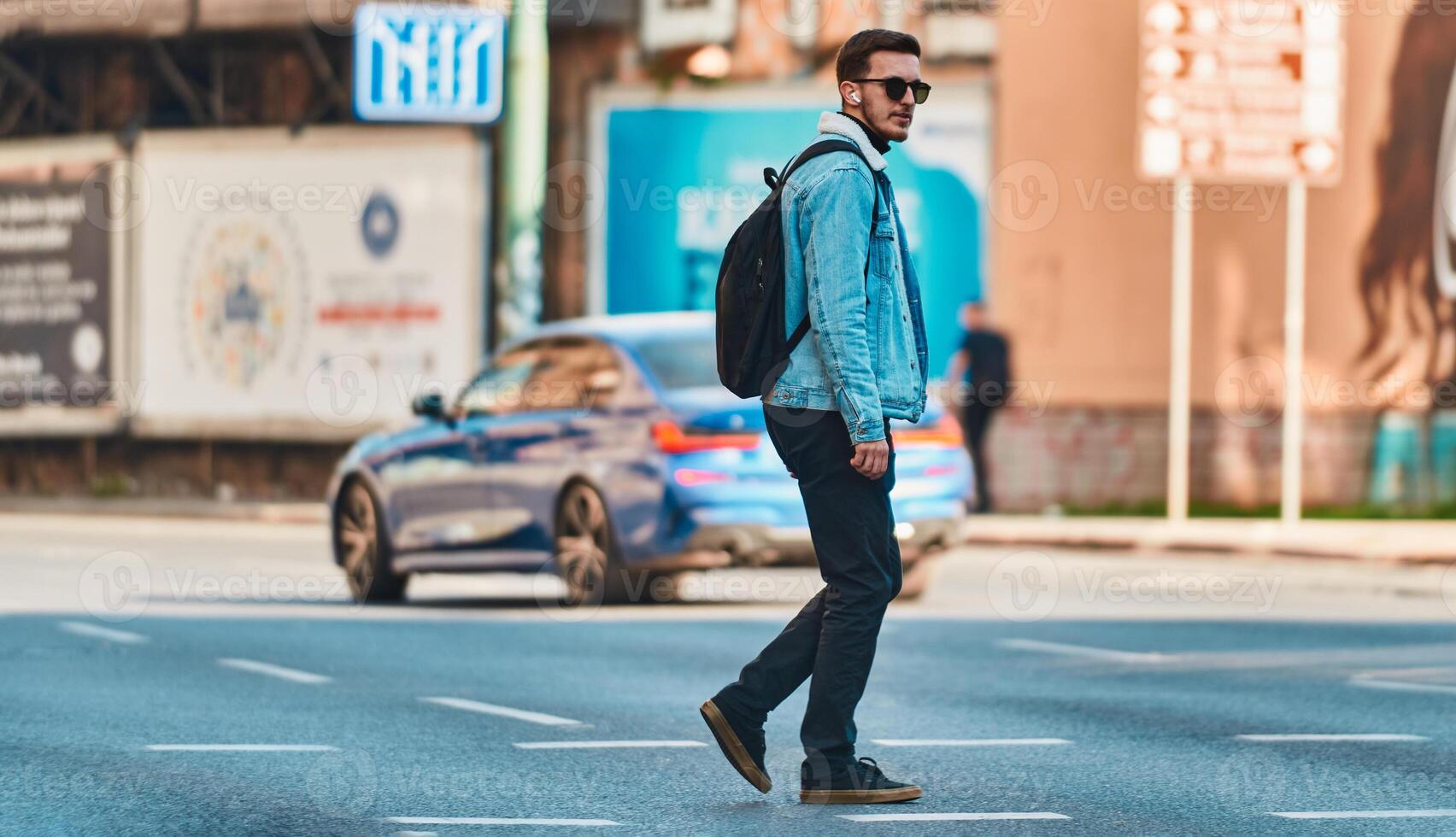 Student walks through the city wearing sunglasses, carrying a backpack, and using wireless headphones, embodying the modern urban lifestyle and the ability to stay connected while on-the-go. photo