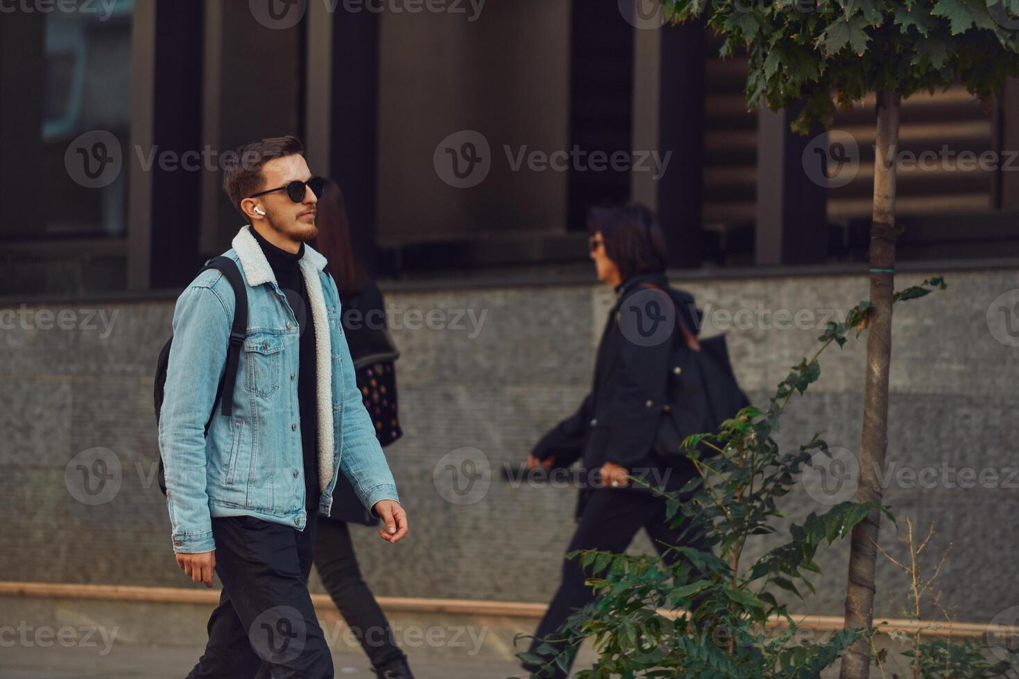 Student walks through the city wearing sunglasses, carrying a backpack, and using wireless headphones, embodying the modern urban lifestyle and the ability to stay connected while on-the-go. photo