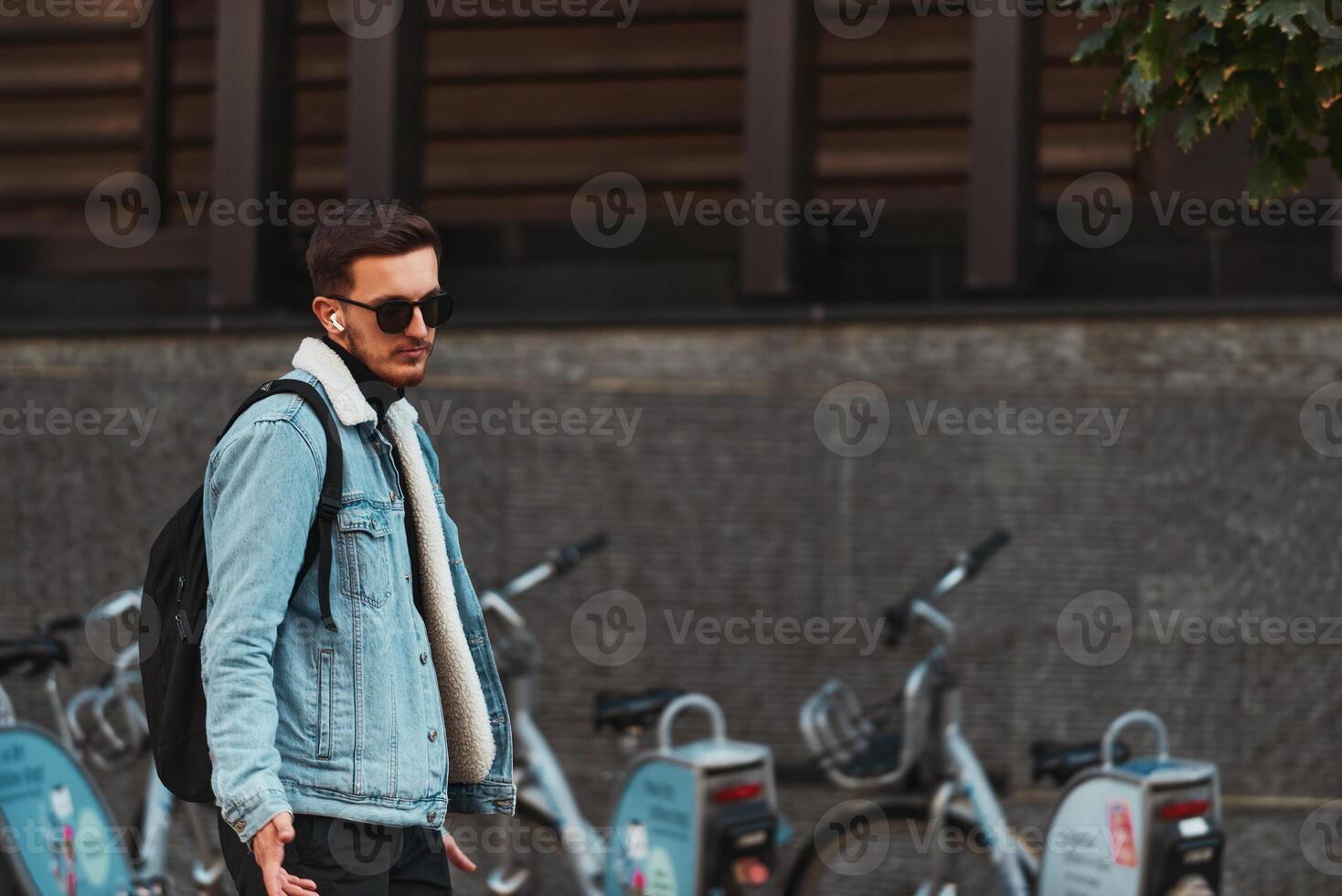 Student walks through the city wearing sunglasses, carrying a backpack, and using wireless headphones, embodying the modern urban lifestyle and the ability to stay connected while on-the-go. photo