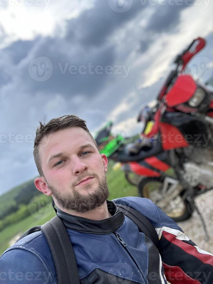 A professional motorcyclist resting on a forest road after an adventurous ride and taking a selfie for social networks photo