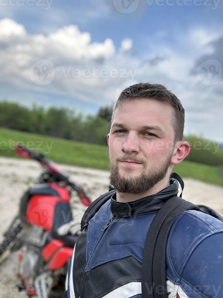 A professional motorcyclist resting on a forest road after an adventurous ride and taking a selfie for social networks photo