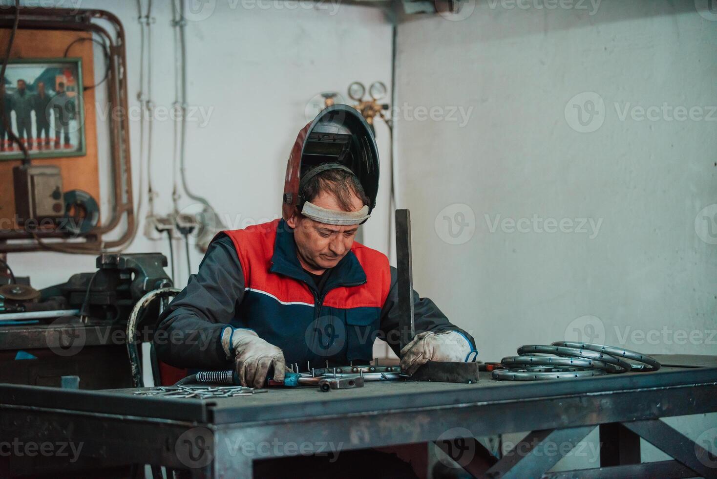 Profesional welder in protective uniform and mask welding metal pipe on the industrial table with other workers behind in the industrial workshop. photo